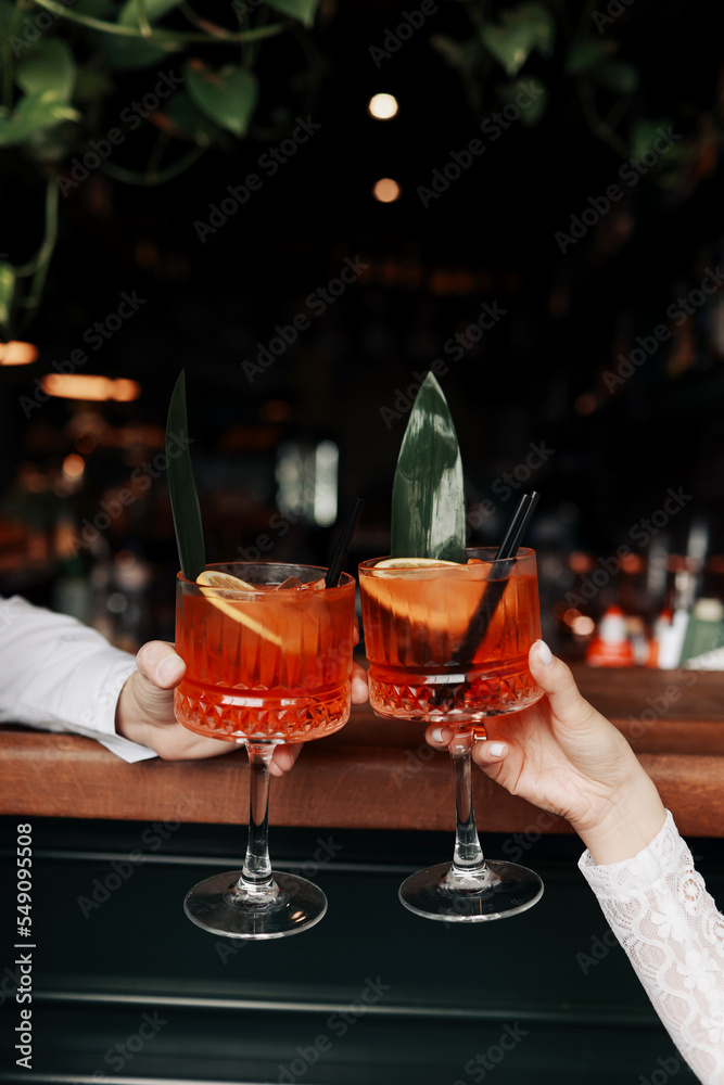 hands of woman and man are clinking, cheers with glasses of Spritz cocktail. Couple celebrating wedding, anniversary with Aperol spritz cocktails, with orange and greens Refreshing alcoholic drink