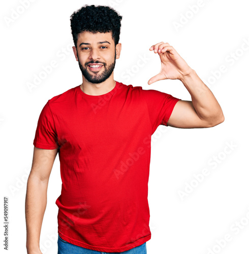 Young arab man with beard wearing casual red t shirt smiling and confident gesturing with hand doing small size sign with fingers looking and the camera. measure concept.