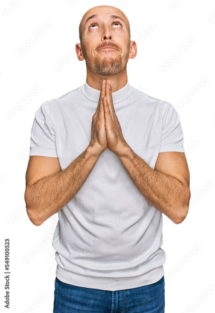 Bald man with beard wearing casual white t shirt begging and praying with hands together with hope expression on face very emotional and worried. begging. Bald man with beard wearing casual white t shirt begging and praying with hands together with hope expression on face very emotional and worried. begging.