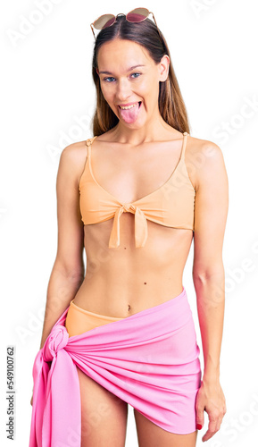Beautiful brunette young woman wearing bikini and sunglasses sticking tongue out happy with funny expression. emotion concept.