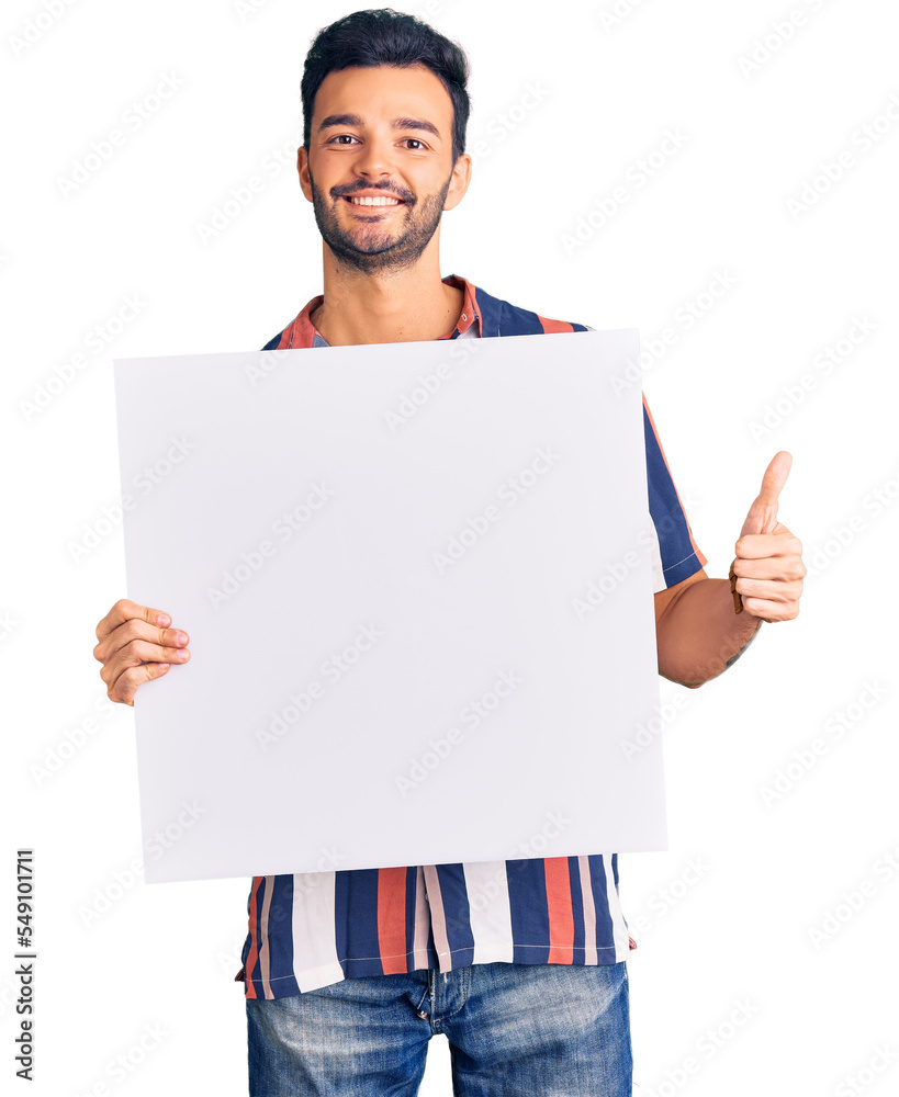 Young handsome hispanic man holding cardboard banner with blank space smiling happy and positive, thumb up doing excellent and approval sign Young handsome hispanic man holding cardboard banner with blank space smiling happy and positive, thumb up doing excellent and approval sign