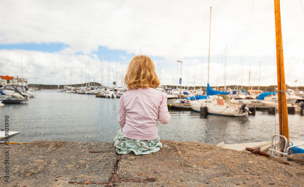 Girl with blond hair sits at the harbor full of boats, child observes yachts and sailboats with interest.
Mädchen mit blonden Haaren sitz am Hafen voller Boote, Kind beobachtet mit Interesse Yachten.