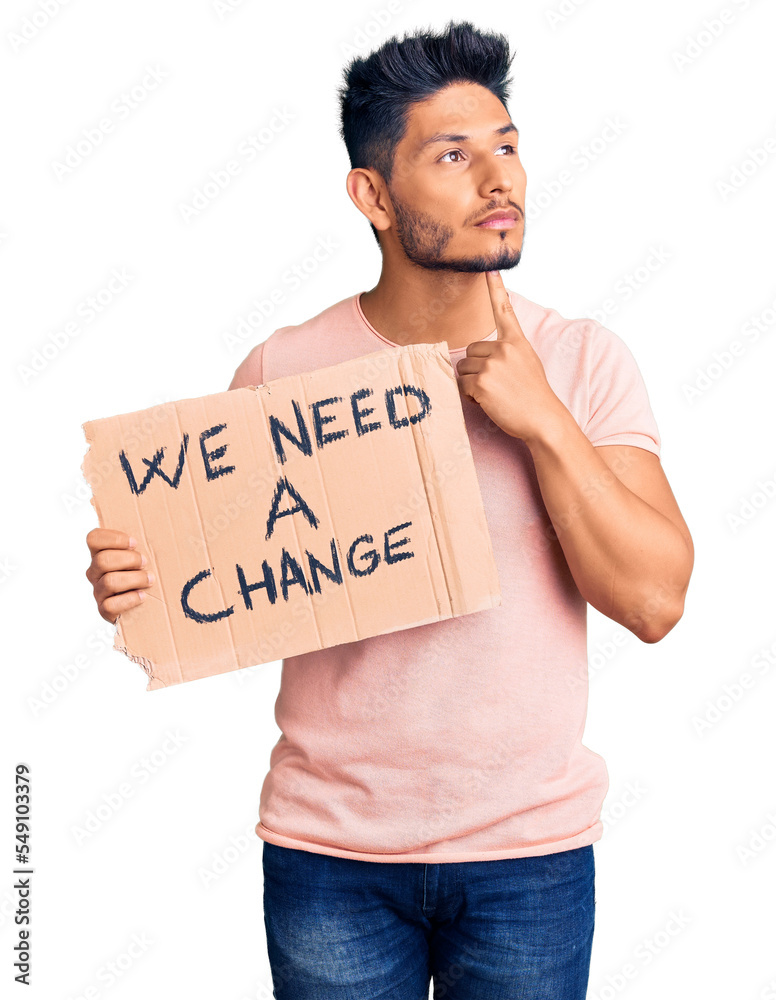 Handsome latin american young man holding we need a change banner serious face thinking about question with hand on chin, thoughtful about confusing idea Handsome latin american young man holding we need a change banner serious face thinking about question with hand on chin, thoughtful about confusing idea