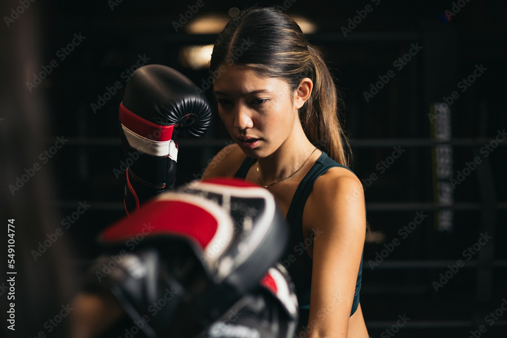 An Asian girl in a boxing ring, the girl trains wearing boxing gloves, the coach greets them with mitts, youth boxing.