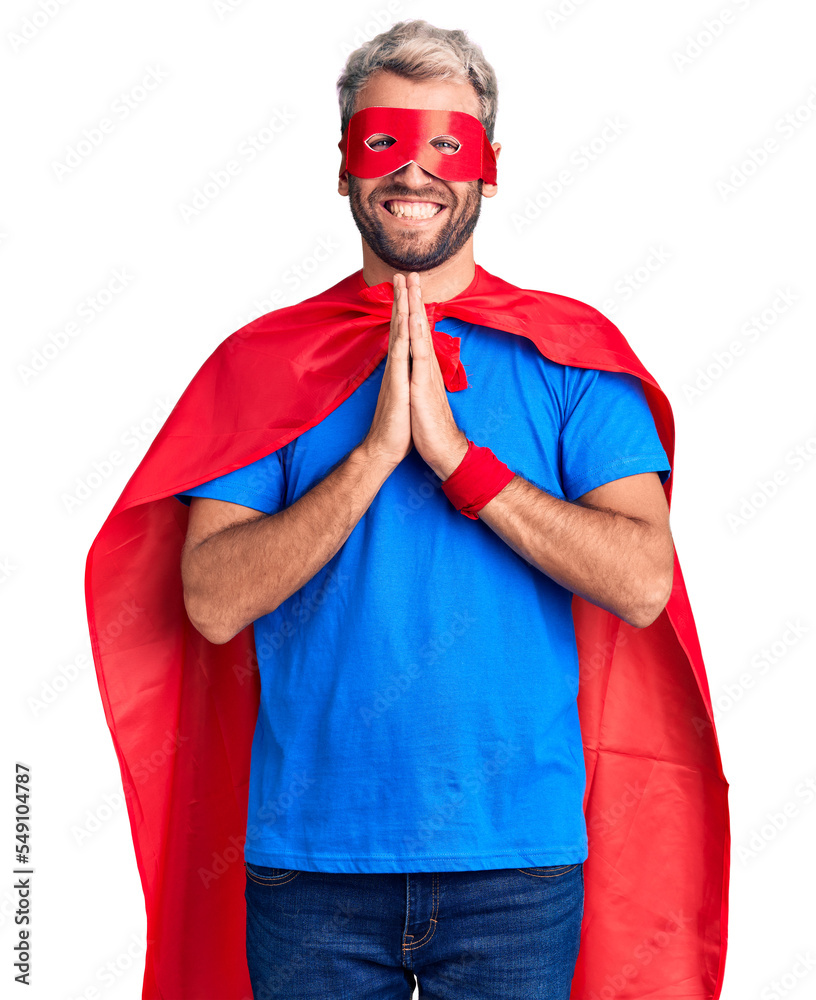 Young blond man wearing super hero custome praying with hands together asking for forgiveness smiling confident. Young blond man wearing super hero custome praying with hands together asking for forgiveness smiling confident.