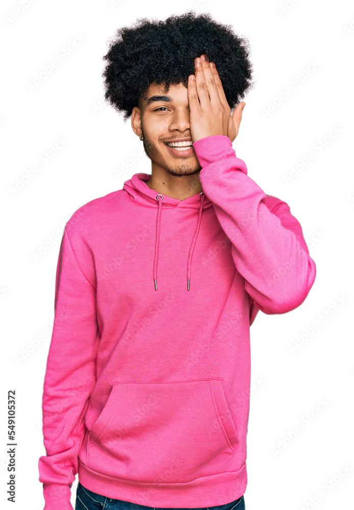 Young african american man with afro hair wearing casual pink sweatshirt covering one eye with hand, confident smile on face and surprise emotion. Young african american man with afro hair wearing casual pink sweatshirt covering one eye with hand, confident smile on face and surprise emotion.