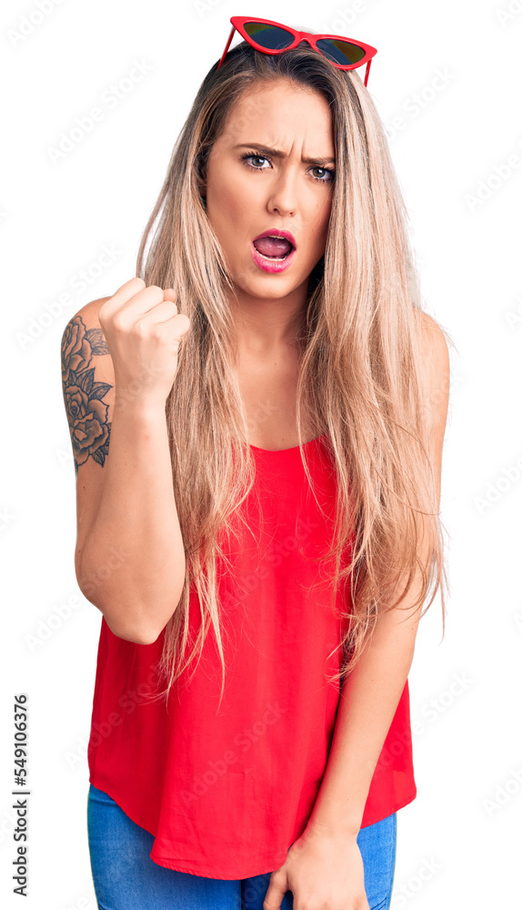 Young beautiful blonde woman wearing sleeveless t-shirt and sunglasses angry and mad raising fist frustrated and furious while shouting with anger. rage and aggressive concept.