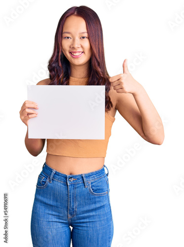 Young beautiful chinese girl holding paper banner with blank space smiling happy and positive, thumb up doing excellent and approval sign