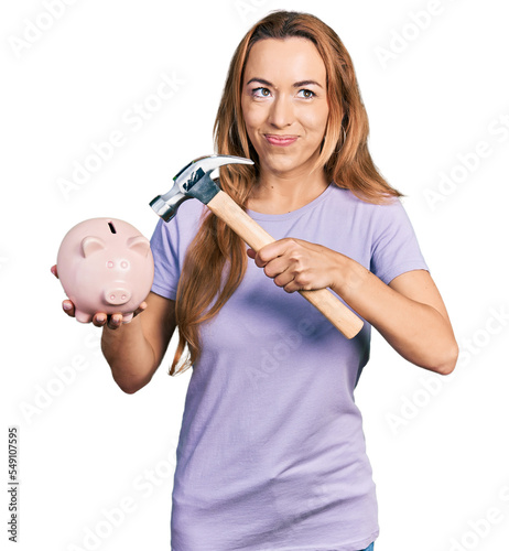Young caucasian woman holding piggy bank and hammer smiling looking to the side and staring away thinking.