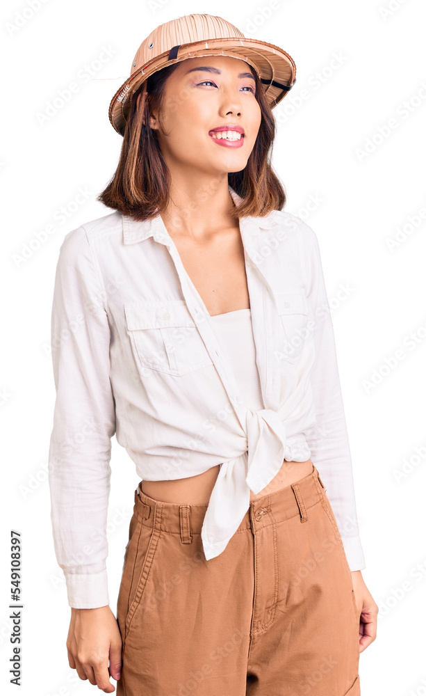 Young beautiful chinese girl wearing explorer hat looking away to side with smile on face, natural expression. laughing confident.