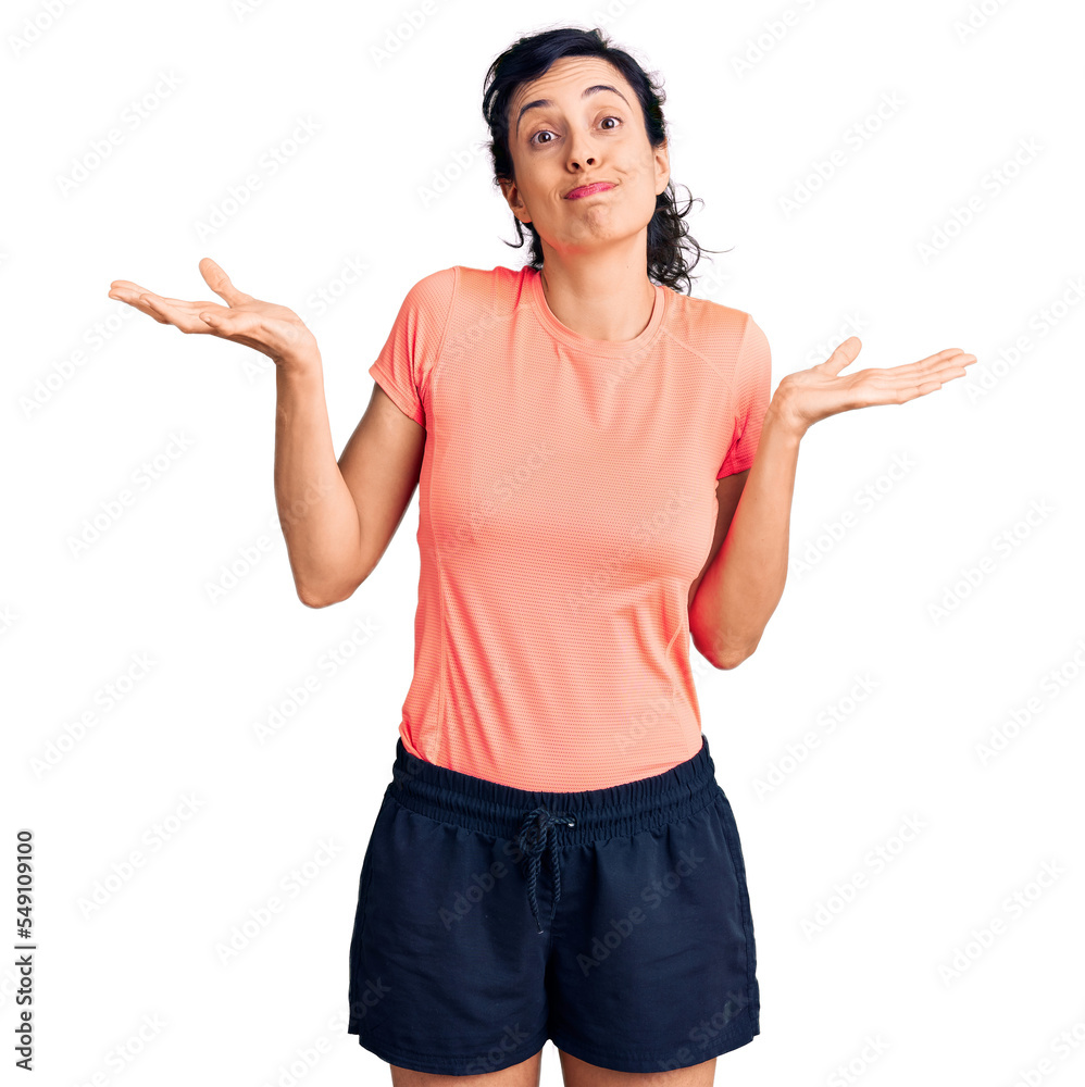 Young beautiful hispanic woman wearing sportswear clueless and confused expression with arms and hands raised. doubt concept. Young beautiful hispanic woman wearing sportswear clueless and confused expression with arms and hands raised. doubt concept.