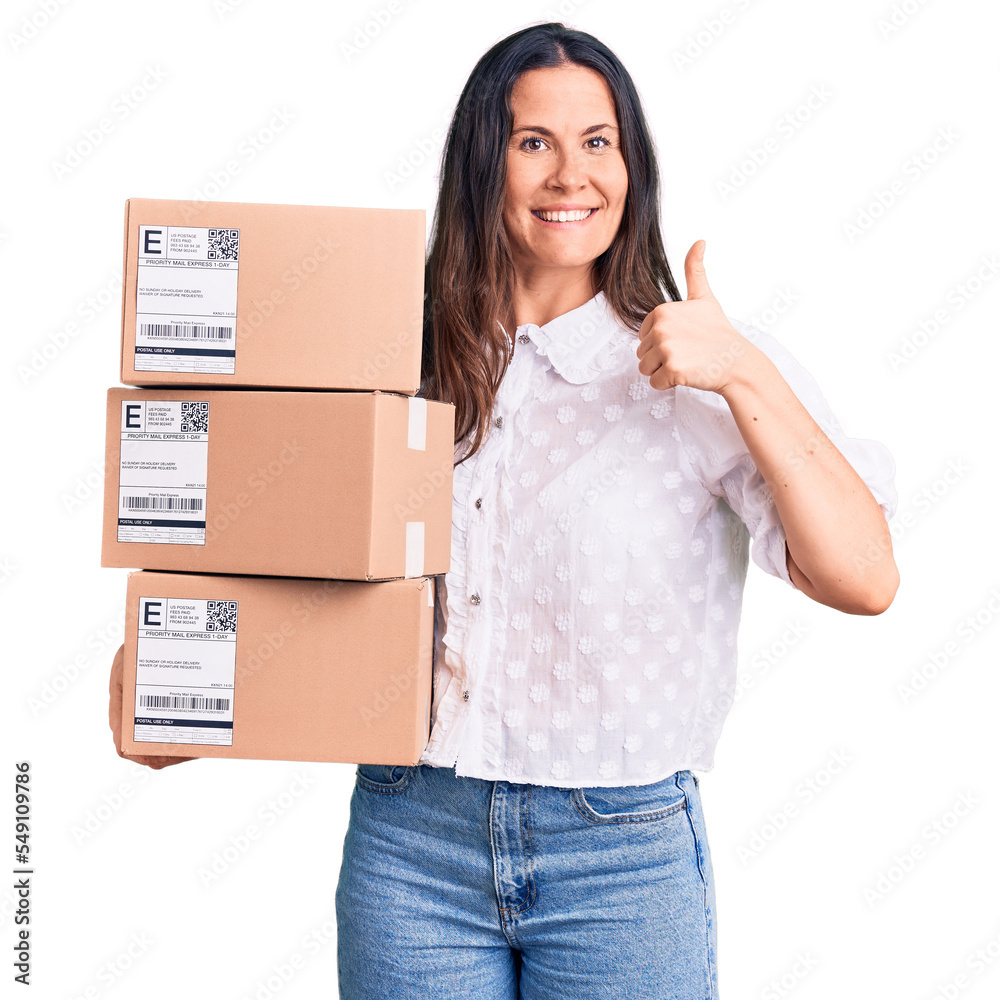 Young beautiful brunette woman holding delivery cardboard package smiling happy and positive, thumb up doing excellent and approval sign Young beautiful brunette woman holding delivery cardboard package smiling happy and positive, thumb up doing excellent and approval sign