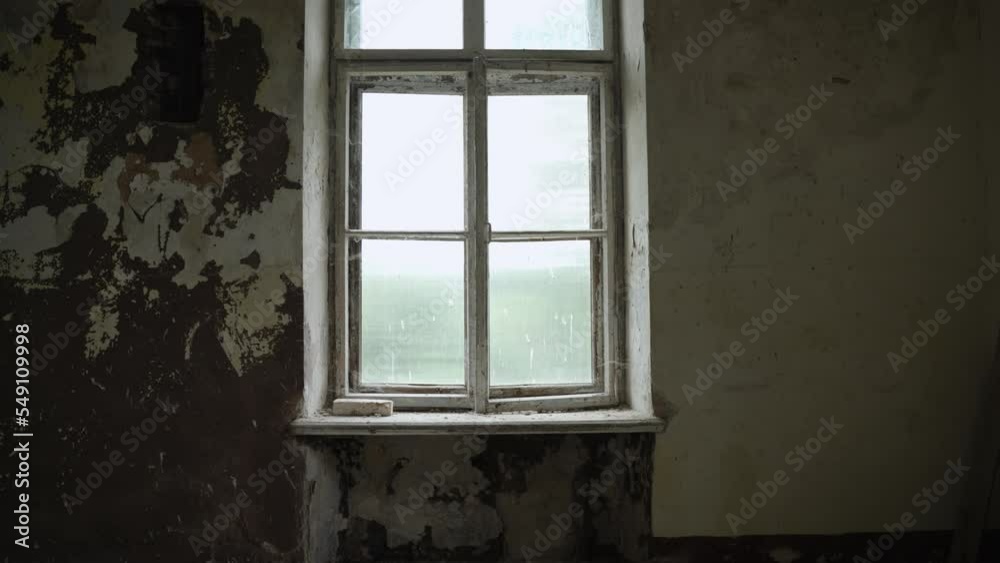 Tall old window with shabby and peeling paint on wooden frame in abandoned empty room, smooth approach of stabilized camera. Dirty window sill overgrown with cobwebs.