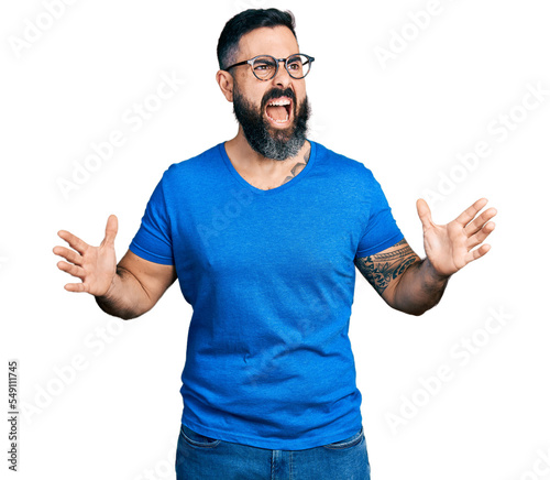 Hispanic man with beard wearing casual t shirt and glasses crazy and mad shouting and yelling with aggressive expression and arms raised. frustration concept. photo