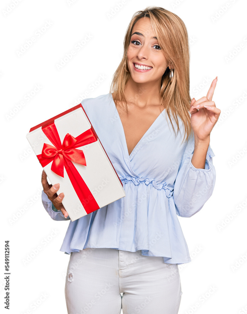 Beautiful blonde woman holding gift surprised with an idea or question pointing finger with happy face, number one Beautiful blonde woman holding gift surprised with an idea or question pointing finger with happy face, number one