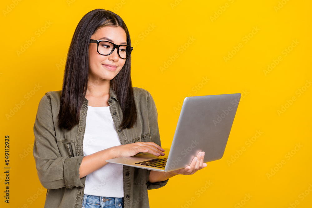 Photo of positive cheerful smart girl with brunette hair wear gray shirt writing email hold laptop isolated on yellow color background Photo of positive cheerful smart girl with brunette hair wear gray shirt writing email hold laptop isolated on yellow color background