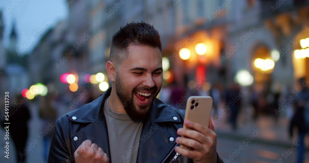 Close up of joyful young handsome happy male in leather jacket videochatting on cellphone and talking emotionally. Outdoor. Attractive guy with beard having video call on smartphone.