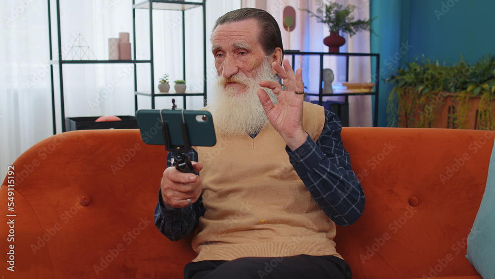 Senior old grandfather man blogger taking selfie on smartphone tripod, communicating, record video call online with subscribers. Elderly grandpa at modern home apartment living room sitting on couch