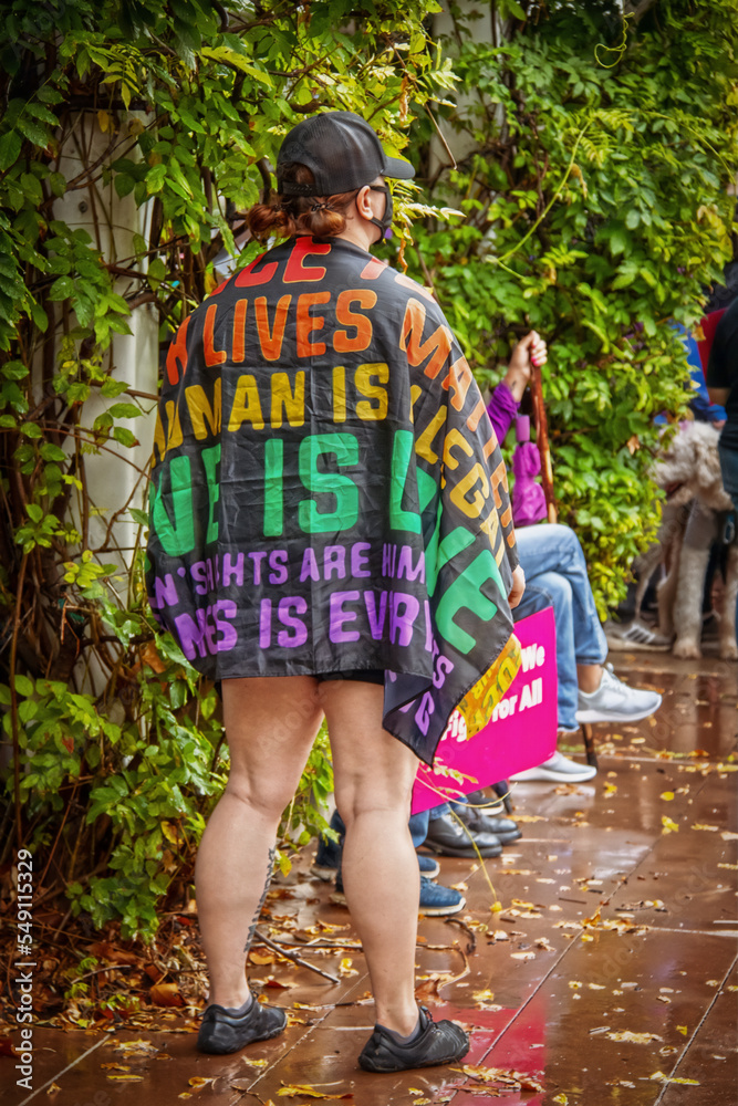 Woman standing at political rally on wet day wrapped in a love is Love All lives matter flag Woman standing at political rally on wet day wrapped in a love is Love All lives matter flag