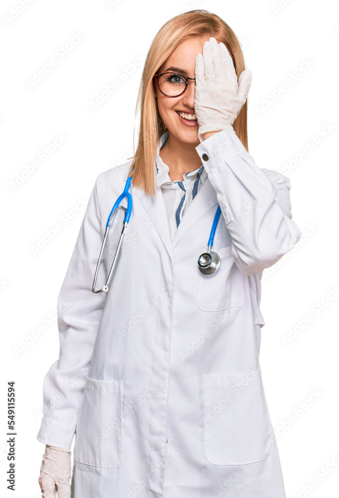 Beautiful caucasian woman wearing doctor uniform and stethoscope covering one eye with hand, confident smile on face and surprise emotion. Beautiful caucasian woman wearing doctor uniform and stethoscope covering one eye with hand, confident smile on face and surprise emotion.
