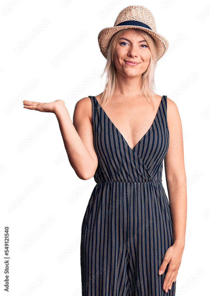 Young beautiful blonde woman wearing summer hat and dress smiling cheerful presenting and pointing with palm of hand looking at the camera. Young beautiful blonde woman wearing summer hat and dress smiling cheerful presenting and pointing with palm of hand looking at the camera.