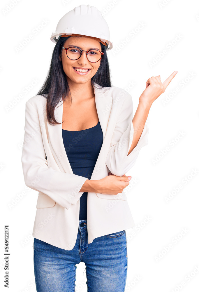 Young beautiful latin girl wearing architect hardhat and glasses with a big smile on face, pointing with hand and finger to the side looking at the camera.