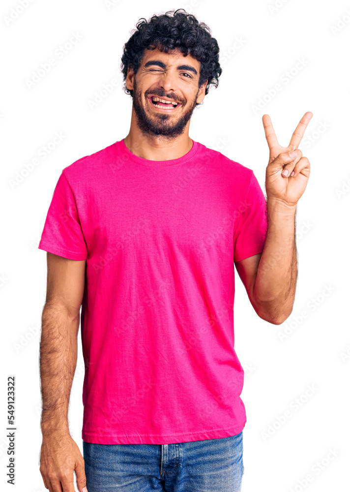 Handsome young man with curly hair and bear wearing casual pink tshirt smiling with happy face winking at the camera doing victory sign. number two. Handsome young man with curly hair and bear wearing casual pink tshirt smiling with happy face winking at the camera doing victory sign. number two.