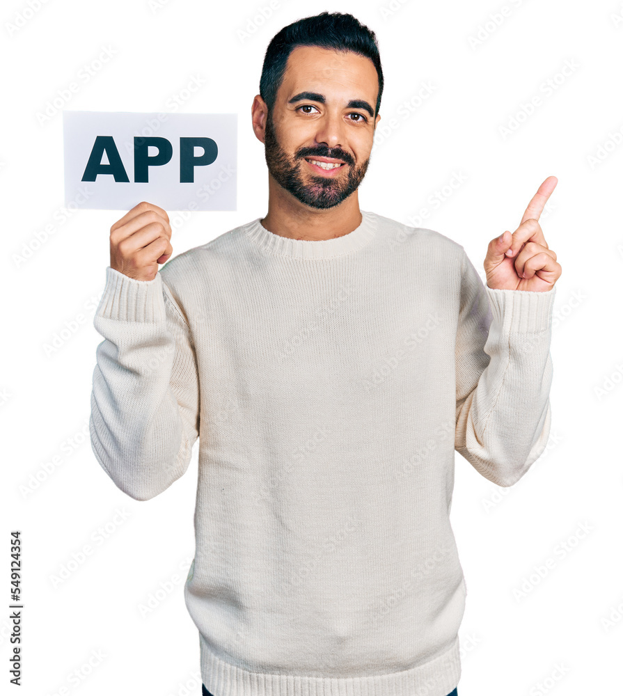 Young hispanic man with beard holding app banner smiling happy pointing with hand and finger to the side Young hispanic man with beard holding app banner smiling happy pointing with hand and finger to the side