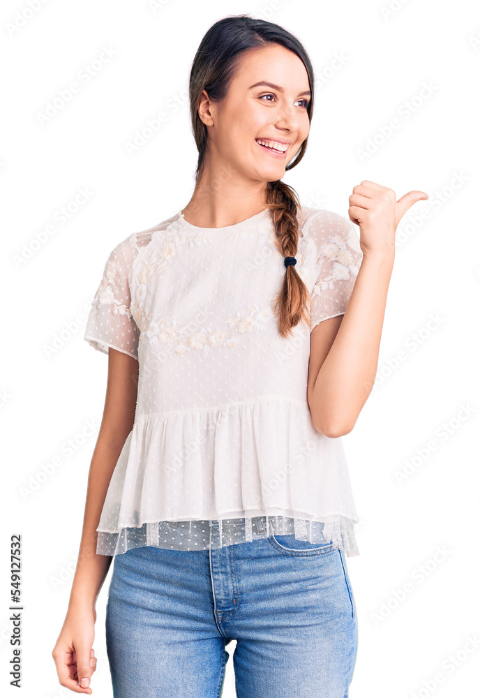Young beautiful girl wearing casual t shirt smiling with happy face looking and pointing to the side with thumb up. Young beautiful girl wearing casual t shirt smiling with happy face looking and pointing to the side with thumb up.