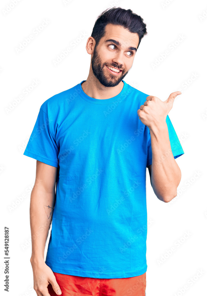 Young handsome man with beard wearing casual t-shirt smiling with happy face looking and pointing to the side with thumb up. Young handsome man with beard wearing casual t-shirt smiling with happy face looking and pointing to the side with thumb up.