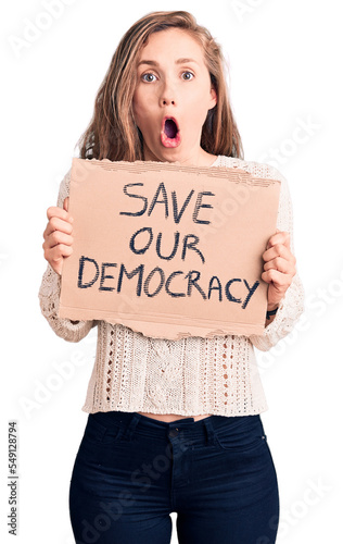Young beautiful blonde woman holding save our democracy banner scared and amazed with open mouth for surprise, disbelief face