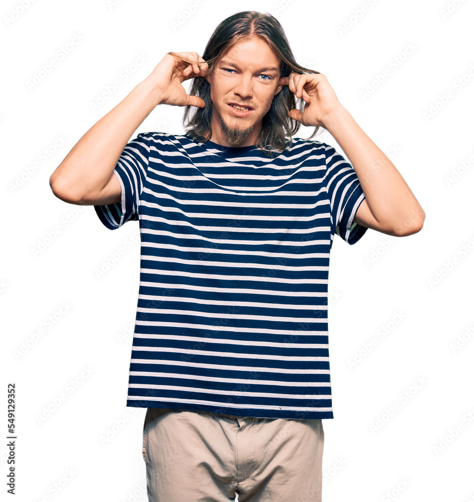 Handsome caucasian man with long hair wearing casual striped t-shirt covering ears with fingers with annoyed expression for the noise of loud music. deaf concept. Handsome caucasian man with long hair wearing casual striped t-shirt covering ears with fingers with annoyed expression for the noise of loud music. deaf concept.