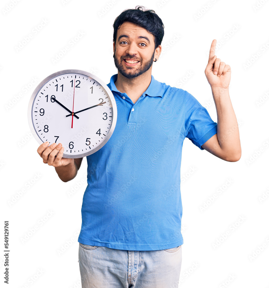 Young hispanic man holding big clock surprised with an idea or question pointing finger with happy face, number one Young hispanic man holding big clock surprised with an idea or question pointing finger with happy face, number one