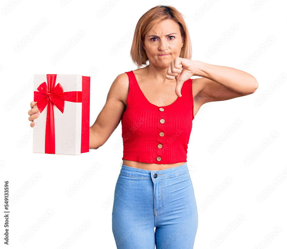 Young blonde woman holding gift with angry face, negative sign showing dislike with thumbs down, rejection concept Young blonde woman holding gift with angry face, negative sign showing dislike with thumbs down, rejection concept