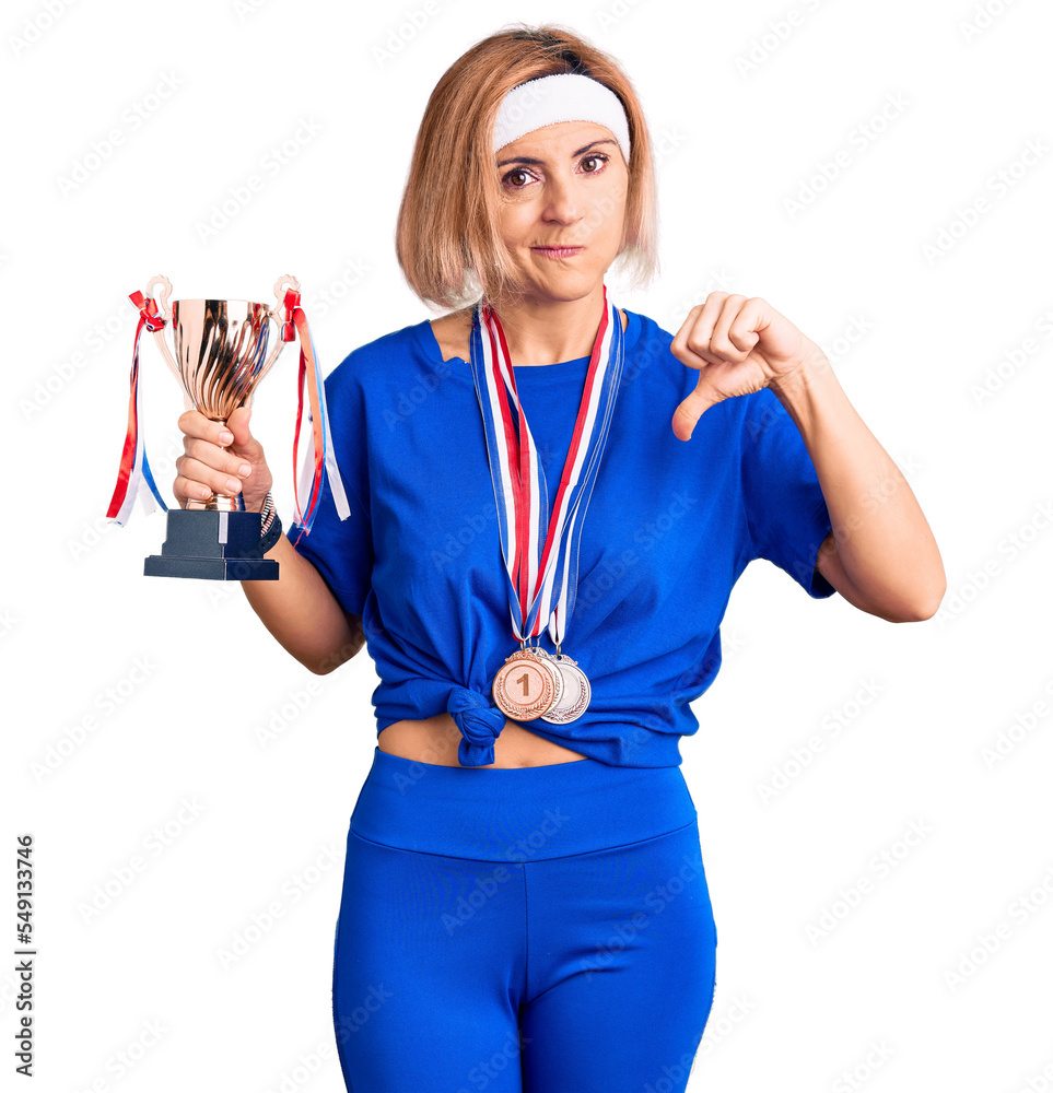Young blonde woman holding champion trophy wearing medals with angry face, negative sign showing dislike with thumbs down, rejection concept
