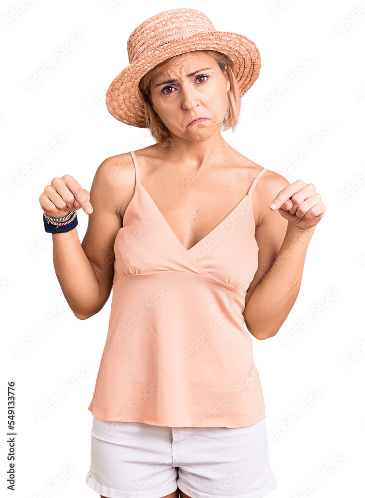 Young blonde woman wearing summer hat pointing down looking sad and upset, indicating direction with fingers, unhappy and depressed. Young blonde woman wearing summer hat pointing down looking sad and upset, indicating direction with fingers, unhappy and depressed.