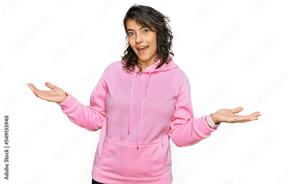 Young hispanic woman wearing casual sweatshirt smiling showing both hands open palms, presenting and advertising comparison and balance Young hispanic woman wearing casual sweatshirt smiling showing both hands open palms, presenting and advertising comparison and balance