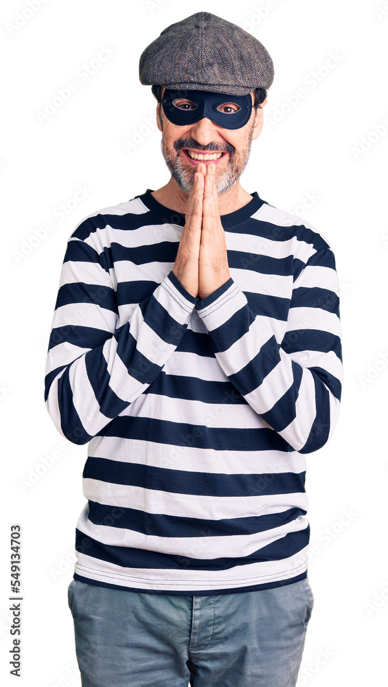 Middle age handsome man wearing burglar mask praying with hands together asking for forgiveness smiling confident. Middle age handsome man wearing burglar mask praying with hands together asking for forgiveness smiling confident.