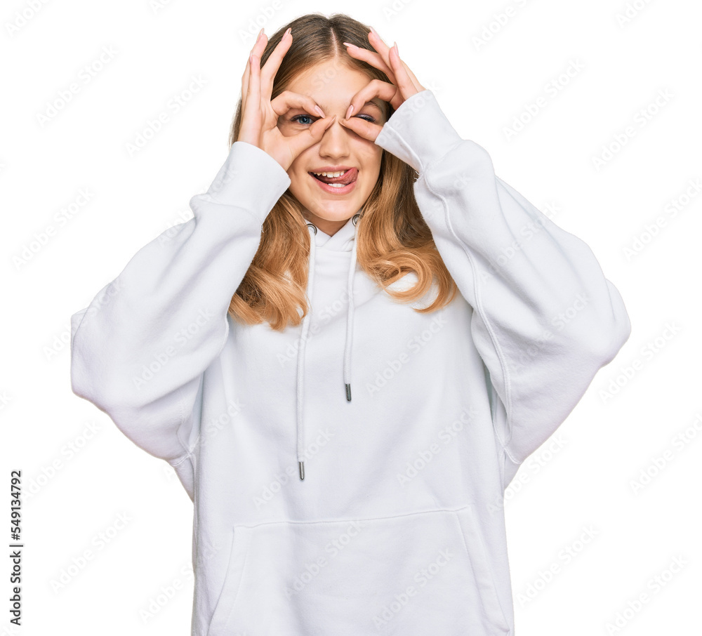 Beautiful young caucasian girl wearing casual sweatshirt doing ok gesture like binoculars sticking tongue out, eyes looking through fingers. crazy expression.
