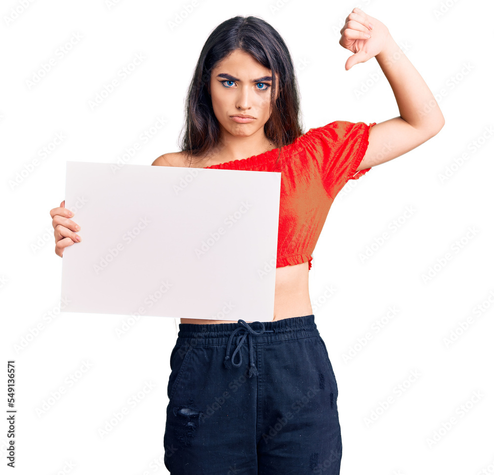 Brunette teenager girl holding blank empty banner with angry face, negative sign showing dislike with thumbs down, rejection concept