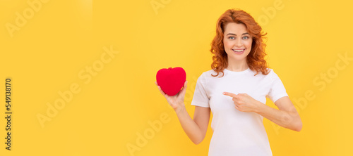 Fotografie, Obraz happy redhead woman pointing finger on love heart on yellow background, love