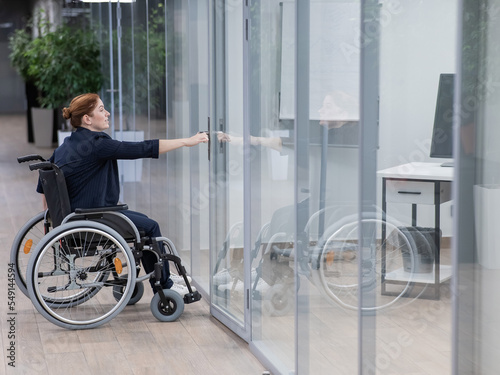 Fototapeta Red-haired caucasian woman in a wheelchair trying to open the door in the office