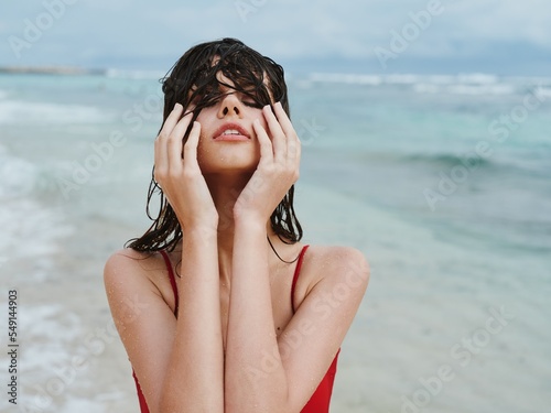 Obraz na plátně Sexy woman in red swimsuit on ocean beach with wet hair covers face with hands,