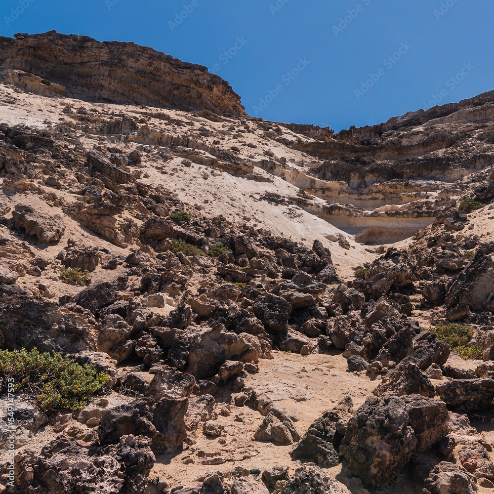 Steep white colored desert cliff with some dark rocks and stones and occasional plants, Fuerteventura 