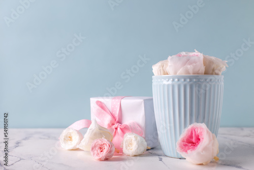omantic background. Postcard with  wrapped box with present and white tender roses flowers in light blue cup against   blue textured  wall.  Place for text. Holiday postcard.
