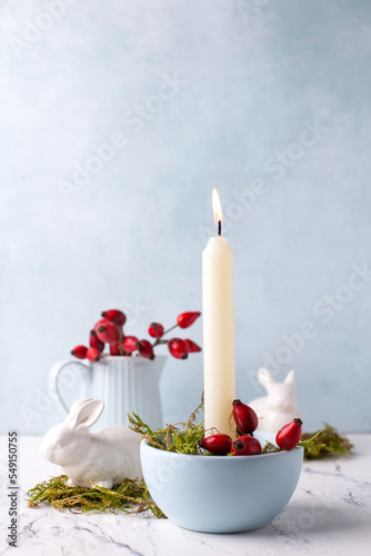 White burning candle, moss, briar berries and white bunnies on white marble background against blue  textured wall. Rabbit is symbool of 2023.. Selective focus. Place for text. photo