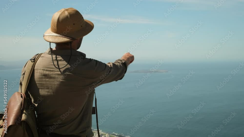 a male traveler in a sun hat points a finger at a famous island in the ocean. hiker man stands on a high mountain against the backdrop of the atlantic ocean. Robben Island, Cape Town, South Africa. a male traveler in a sun hat points a finger at a famous island in the ocean. hiker man stands on a high mountain against the backdrop of the atlantic ocean. Robben Island, Cape Town, South Africa.