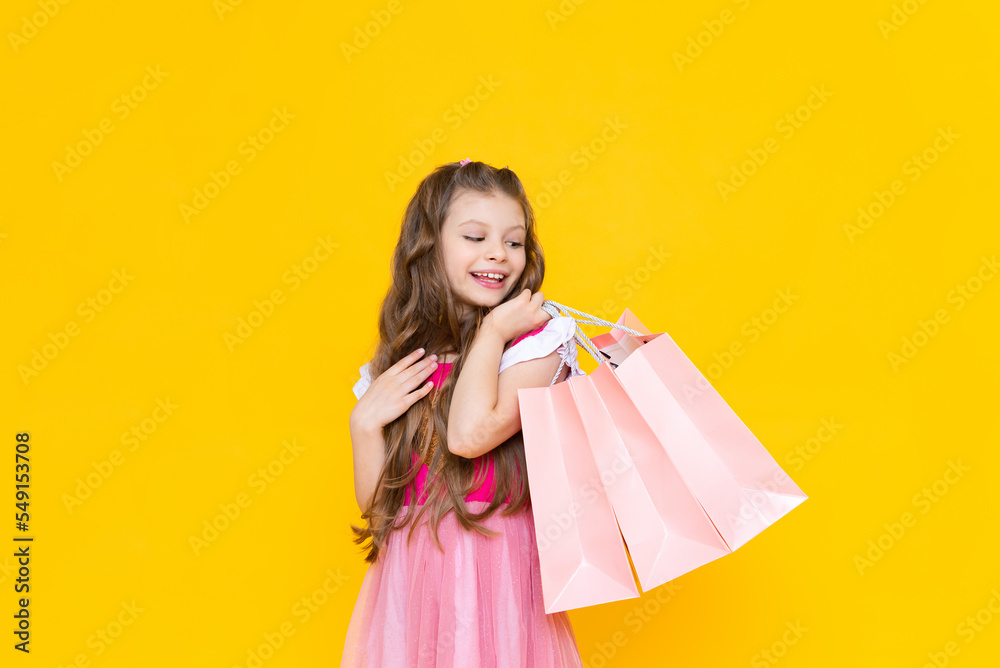 A child holds paper bags with purchases after shopping. Sale and shopping for little girls. A charming girl holds gift bags on a yellow isolated background.