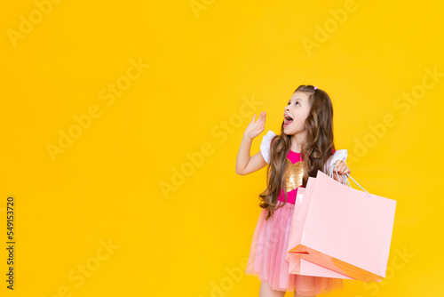 A child holds paper bags with purchases after shopping. Sale and shopping for little girls. A charming girl holds gift bags on a yellow isolated background. Copy space.