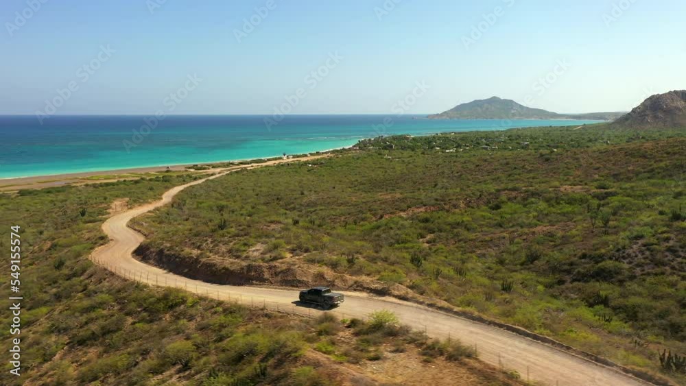 Aerial Panning Shot Of Car Moving On Road By Turquoise Sea Against Clear Sky - Baja California, Mexico Aerial Panning Shot Of Car Moving On Road By Turquoise Sea Against Clear Sky - Baja California, Mexico